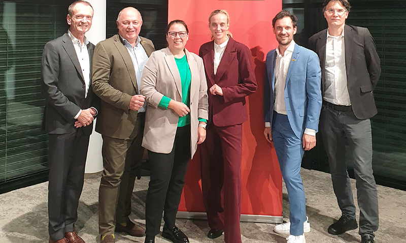 Sechs Personen stehen nebeneinander vor einem roten Hintergrund, alle in formeller Kleidung. Zwei tragen Anzüge in Braun- und Blautönen, während eine Person einen auffälligen roten Anzug trägt. Die Gruppe posiert lächelnd für das Foto.