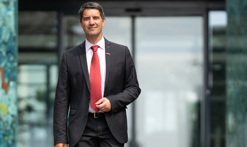 LK im Portrait: Ralf Mittermayr / Saubermacher Mann im schwarzen Anzug mit roter Krawatte vor einem modernen Bürogebäude mit Glastüren
