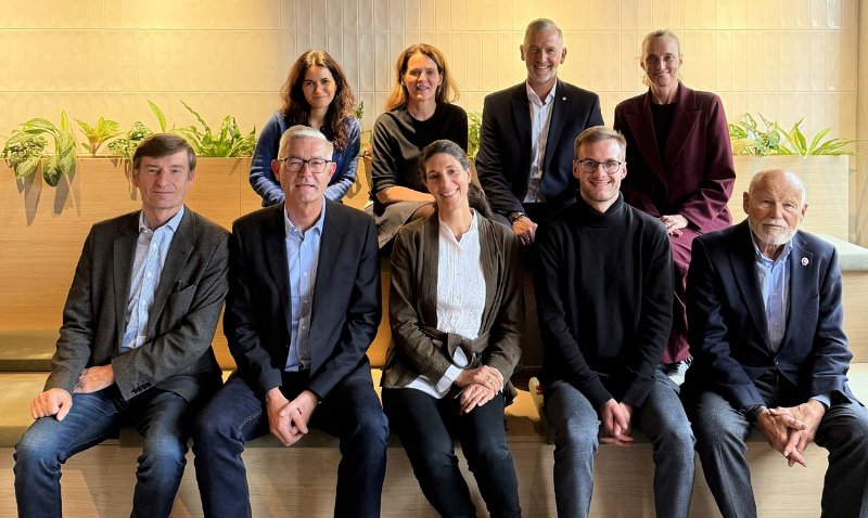 Eine Gruppe von neun Personen sitzt und steht in zwei Reihen vor einer Holzwand mit Pflanzen. Die vordere Reihe sitzt auf einer Bank, während die hintere Reihe steht. Alle tragen formelle oder semi-formelle Kleidung.