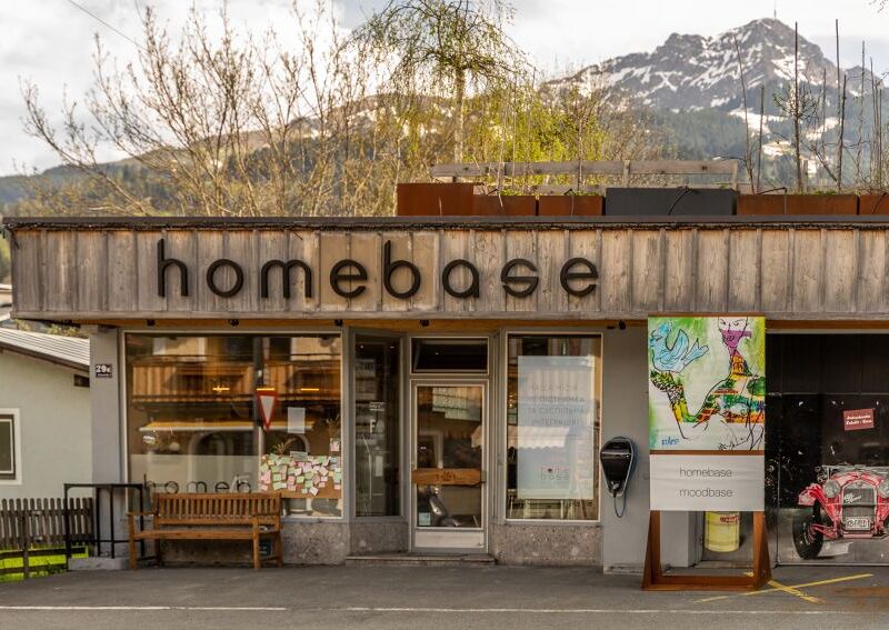 Außenansicht eines Geschäfts mit dem Namen 'homebase' in einer malerischen Berglandschaft, mit einem Holzbänkchen und einer bunten Karte neben dem Eingang.
